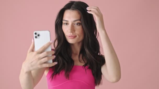一位漂亮的年轻女子正在用她的智能手机拍摄自己的照片 她站在粉色的背景下 与外界隔绝 — 图库视频影像