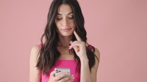 笑顔の若い女性がピンクの背景に隔離されたスマートフォンに入力しています — ストック動画
