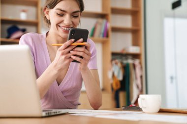 Ofiste dizüstü bilgisayarla çalışırken cep telefonu kullanan gülümseyen bir kadının fotoğrafı.