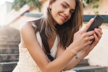 Pozitif iyimser bir genç kadının cep telefonu kullanırken merdivenlerde oturmasının fotoğrafı..