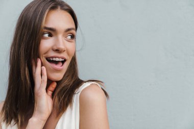 Dışarı bakarken gri duvarın üzerinde poz veren güzel, şaşkın, iyimser genç bir kadın görüntüsü..