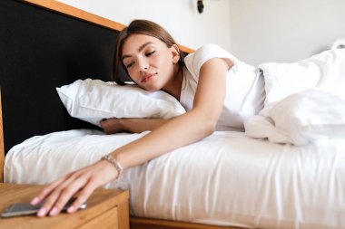 Çekici genç bir kadın sabah yatakta uzanırken cep telefonu kullanıyor, telefona uzanıyor.