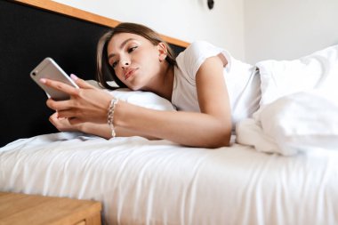 Sabahları yatakta uzanırken cep telefonu kullanan çekici genç bir kadın.