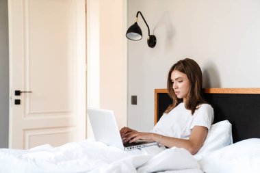 Yatakta dinlenirken dizüstü bilgisayar kullanan çekici genç bir kadın.