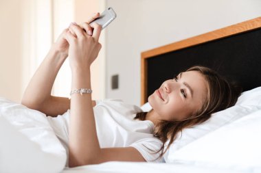 Sabahları yatakta uzanırken cep telefonu kullanan çekici, gülümseyen genç bir kadın.