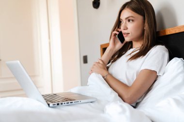 Yatakta dinlenirken cep telefonu ve dizüstü bilgisayar kullanan çekici genç bir kadın.