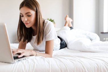 Yatakta dinlenirken dizüstü bilgisayar kullanan çekici genç bir kadın.