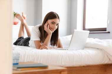 Yatakta dinlenirken dizüstü bilgisayar kullanan çekici genç bir kadın.