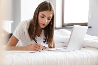 Güzel genç bir kadın dizüstü bilgisayarla yatakta yatarken not alıyor.