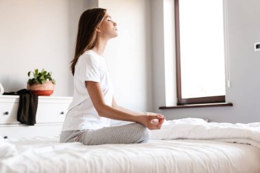 Genç bir kadın, parlak yatak odasında gözleri kapalı meditasyon yapıyor, yatakta oturuyor.