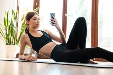 Formda spor yapan genç bir kadın evde spor paspası üzerinde egzersiz yaparken cep telefonu kullanıyor..