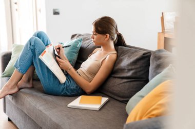 Evdeki kanepede oturmuş notlar alan çekici genç bir kadın.