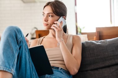 Evdeki kanepede dizüstü bilgisayar ve cep telefonu kullanan sıradan bir genç kadın.