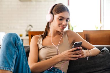 Evde kulaklıkla müzik dinleyen, elinde cep telefonu olan rahat bir kadın.