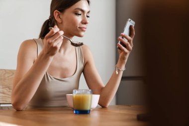 Evdeki masada kahvaltı ederken cep telefonuyla konuşan güzel genç bir kadın.