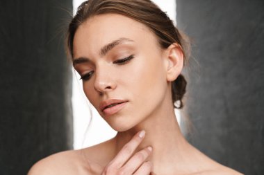 Gri arka planda tek başına duran güzel, beyaz tenli, üstsüz bir kadın resmi.