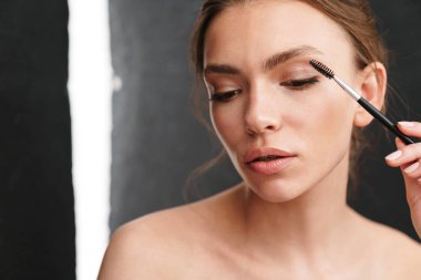 Gri arkaplandan izole edilmiş fırçayla makyaj yapan beyaz, genç, güzel bir kadın resmi