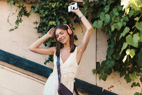 一个漂亮的年轻女子在户外跳舞 一边用耳机听音乐 一边拿着手机的照片 — 图库照片