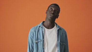 Sıkılmış bir Afrikalı Amerikalı genç adam portakal rengi bir arka planda tek başına kameraya bakıyor.