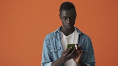 Odaklanmış bir Afrikalı Amerikalı genç, turuncu arka planda tek başına duran akıllı telefonuyla yazı yazıyor.