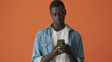 Yakışıklı bir Afrikalı Amerikalı genç adam akıllı telefonunu turuncu arka planda tek başına duruyor.