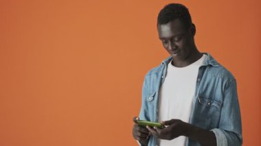 Çekici bir Afrikalı Amerikalı genç adam akıllı telefonuyla bir şey izliyor turuncu arka planda yatay olarak duruyor.