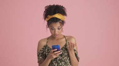 Şaşırmış bir Afrikalı Amerikalı genç kadın, pembe arka planda tek başına duran akıllı telefonunu kullanarak inanılmaz haberler alıyor.