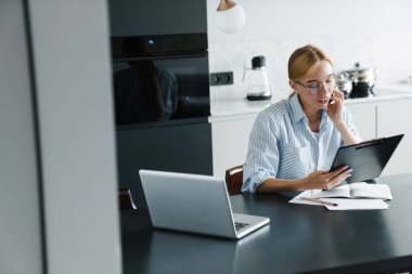 Mutfaktaki masada dizüstü bilgisayarla çalışırken cep telefonuyla konuşan gözlüklü ciddi bir genç kadının fotoğrafı.