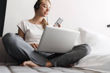 Güzel bir genç kadının cep telefonu ve kulaklık kullanırken parlak odada dizüstü bilgisayarla çalışırken çekilmiş fotoğrafı.