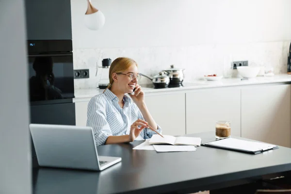 여성이 부엌에 책상에서 노트북 컴퓨터로 일하면서 휴대폰으로 이야기하는 — 스톡 사진