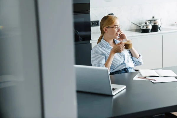 여성이 부엌에서 노트북 컴퓨터로 일하는 커피를 마시고 핸드폰으로 이야기하며 — 스톡 사진