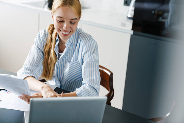 Photo of happy young woman working with papers and laptop while sitting at table in kitchen