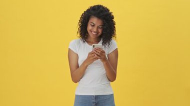 Sarı duvar arkasında izole edilmiş cep telefonu kullanan, gülümseyen genç Afrikalı kadın.