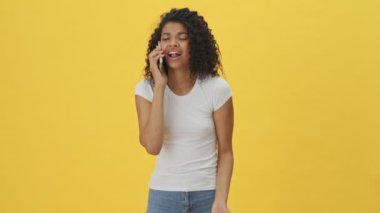 Sarı duvar arkasında izole edilmiş cep telefonuyla konuşan genç Afrikalı mutlu kadın.
