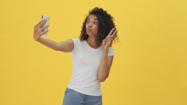 Genç Afrikalı bir kadın cep telefonuyla selfie çekiyor. Sarı duvar arkasında izole edilmiş.