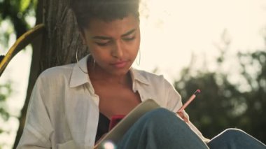 Güzel, Afro-Amerikan bir kadın parktaki çimlerin üzerine oturmuş notlar alıyor.