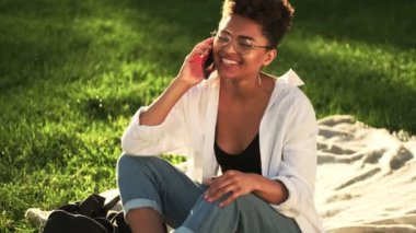Gözlüklü, gülen Afro-Amerikan bir kadın parkta piknik saatinde çimlerin üzerinde otururken akıllı telefonuyla konuşuyor.
