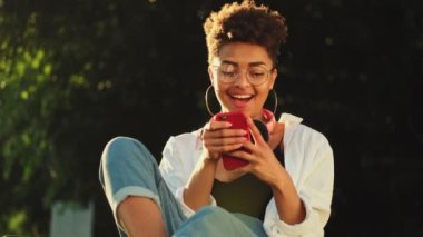 Gözlüklü, gülümseyen Afro-Amerikan bir kadın parkta piknik saatinde çimlerin üzerinde otururken akıllı telefonunu kullanıyor.
