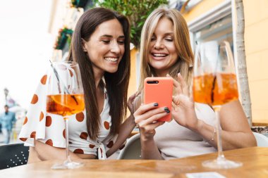 Neşeli yetişkin görüntüsü. İki kadın gülümsüyor ve cep telefonu kullanıyor. Açık kafedeki masada kokteyl içerken.
