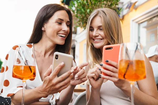 Kafede kokteyl içerken gülümseyen ve cep telefonu kullanan iki kadının görüntüsü.