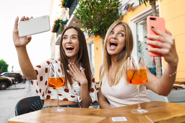 图为两名兴奋的成年女性在户外咖啡桌上喝鸡尾酒时 在手机上自拍的照片 — 图库照片
