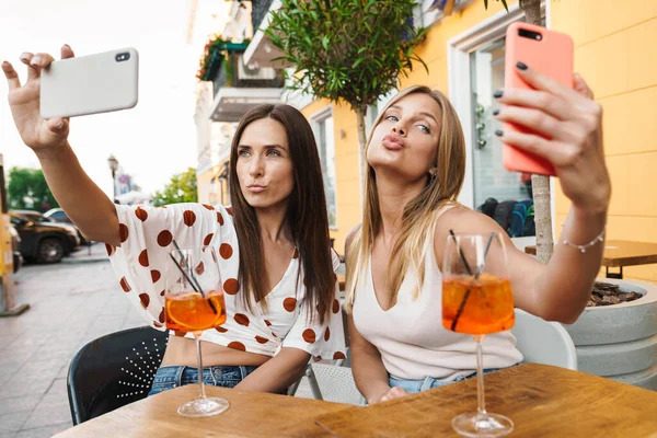 图为两个快乐的成年女子在户外咖啡桌上喝鸡尾酒时 在手机上自拍的情景 — 图库照片