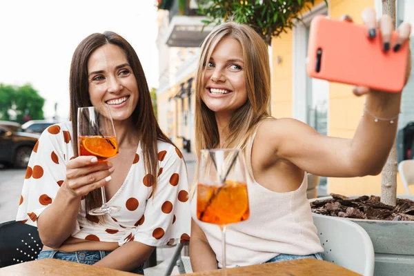 Neşeli yetişkin iki kadının cep telefonuyla fotoğraf çekerken açık kafedeki masada kokteyl içerkenki görüntüsü.