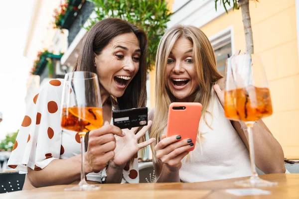 Dışarıdaki kafede kokteyl içerken ellerinde kredi kartı ve cep telefonu olan heyecanlı iki kadının görüntüsü.
