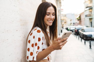 Şehirde yürürken gülümseyen ve cep telefonu kullanan mutlu esmer kadın görüntüsü