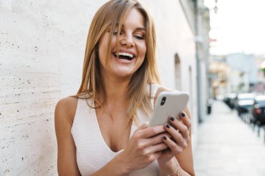 Şehir caddesinde yürürken gülen ve cep telefonu kullanan mutlu sarışın kadın görüntüsü