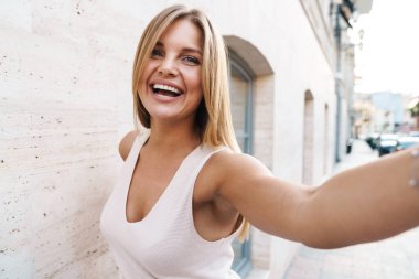 Şehirde yürürken gülümseyen ve selfie çeken neşeli beyaz kadın görüntüsü