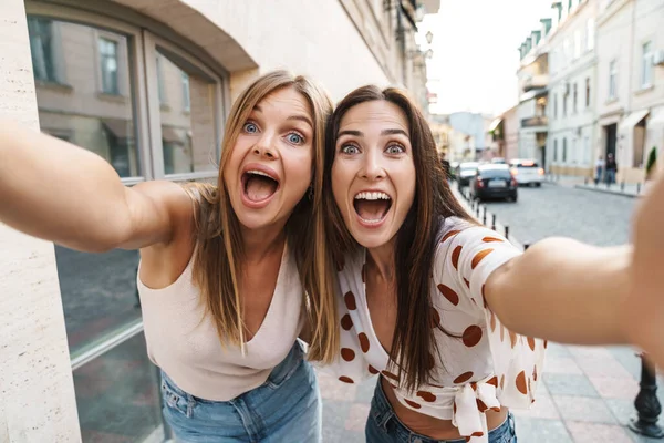 Image of excited adult two women hugging and taking selfie photo while walking on city street