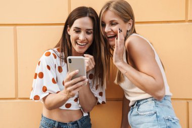 Neşeli yetişkin iki kadının şehir sokaklarında poz verirken cep telefonu kullanması ve gülmesi.