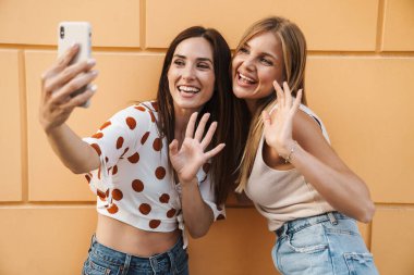 Neşeli yetişkin iki kadının cep telefonuyla selfie çekmesi ve şehirde poz verirken el sallaması.
